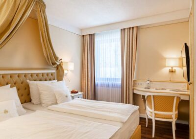 Hotelzimmer mit zwei Einzelbetten, gepolstertem Kopfteil mit Baldachin, Schreibtisch mit Stuhl, Wandfernseher und großem Fenster mit Vorhang.
